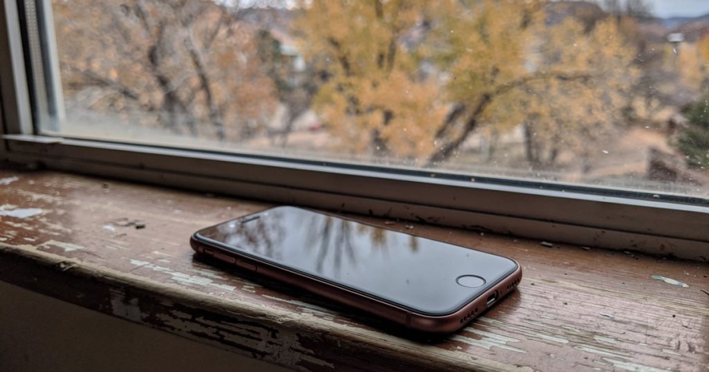A smartphone on a kitchen windowsill with autumn cottonwood trees visible through the glass in morning light