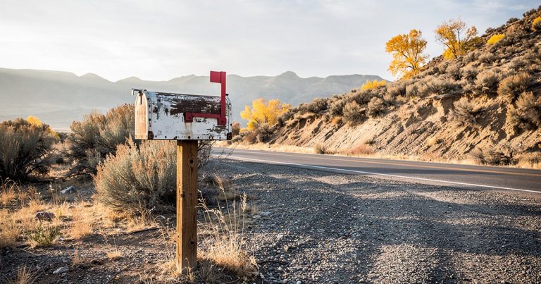A weathered rural mailbox with its flag raised on a gravel verge, autumn cottonwoods and Utah foothills in the background under a pale morning sky