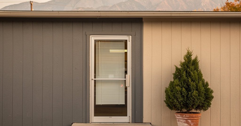 Exterior of a small community clinic building on a quiet Utah morning, Wasatch foothills in the distance under a pale autumn sky